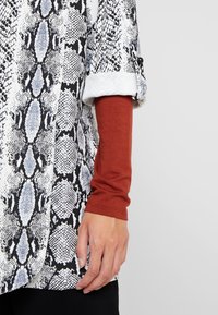 Een overhemd met slangenprint in zwart, wit en grijs met opgerolde mouwen, gecombineerd met een aansluitende armwarmer in roestkleur.