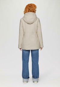 Beige gewatteerde jas met een getailleerde pasvorm, zichtbare stikdetails, gecombineerd met een paar wijde blauwe jeans. De stof van de jas lijkt gestructureerd.