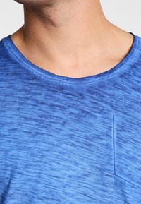 Niebieska koszulka z okrągłym dekoltem, z teksturowanym splotem. Widoczna jest mała kieszeń, podkreślona kontrastowym przeszyciem.