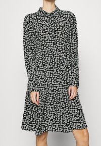 Abito nero con maniche lunghe, colletto e orlo a strati. Presenta un motivo floreale bianco su un tessuto leggero.