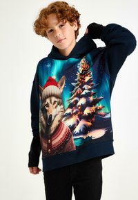 Hoodie met een wolf die een rode hoed en een feestelijk sweater draagt, tegen een kleurrijk winterlandschap met een kerstboom.