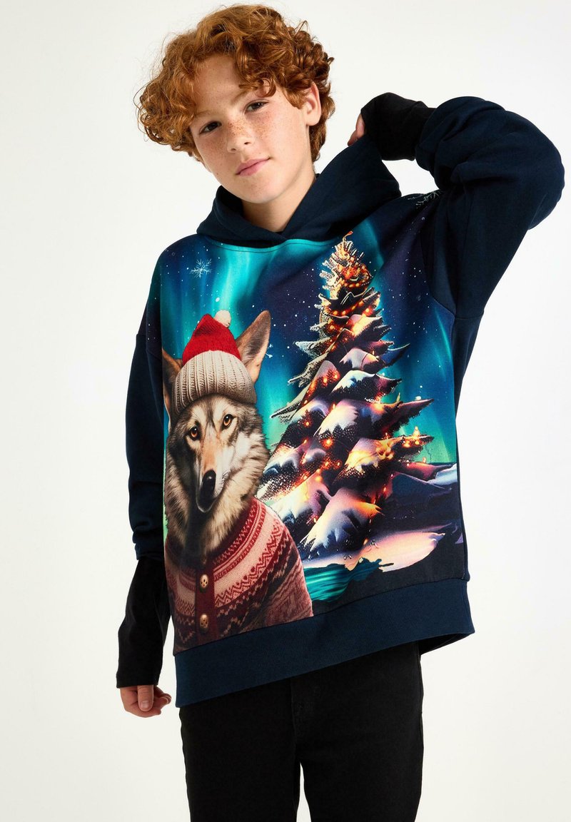 Hoodie met een wolf die een rode hoed en een feestelijk sweater draagt, tegen een kleurrijk winterlandschap met een kerstboom.