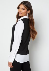 Top nero senza maniche sopra una camicia bianca a maniche lunghe, con orlo arrotondato, tessuto liscio e forma aderente, abbinato a pantaloni neri.