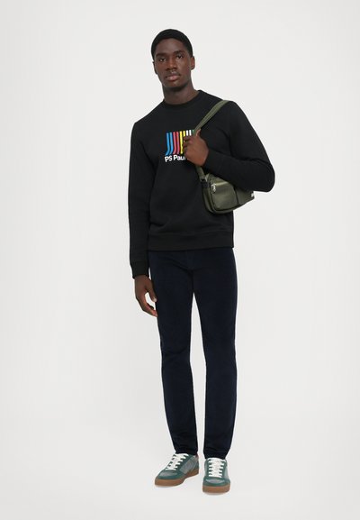 Svart hettegenser med fargerike striper og "PS Paul"-logo, mørkeblå bukser, grønne sneakers med hvite snorer, og en grønn crossbody-veske.