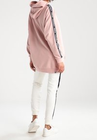 Rosa huvtröja med svarta sidostripe som har "PUMA"-logotyp. Kombinerad med vita joggers som visar en avslappnad passform och mjukt material.