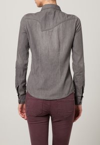 Maglietta grigia a maniche lunghe con orlo curvo, colletto e polsini abbottonati, abbinata a pantaloni bordeaux con tasche posteriori. Tessuto morbido, simile al denim.