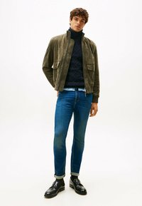 Giovane uomo che indossa una giacca di camoscio oliva, un maglione a collo alto blu navy, jeans blu con risvolti e mocassini di pelle nera, in piedi su uno sfondo bianco.