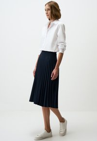 Marineblauwe geplooide midi-rok van lichtgewicht stof, gecombineerd met een witte blouse met kraag en witte sneakers, op een effen achtergrond.