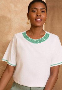 Haut blanc à manches courtes avec un col et des bordures perlés verts. Tissu doux avec une coupe décontractée et une texture subtile.