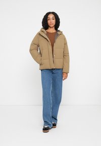 Giacca puffer color beige con cappuccio, caratterizzata da un design trapuntato e chiusure a pressione, abbinata a jeans a gamba larga blu chiaro e scarpe nere.