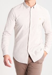 Camisa de manga larga beige claro con botones y una textura sutil, que presenta un pequeño logo dorado en el pecho y cuello estándar.