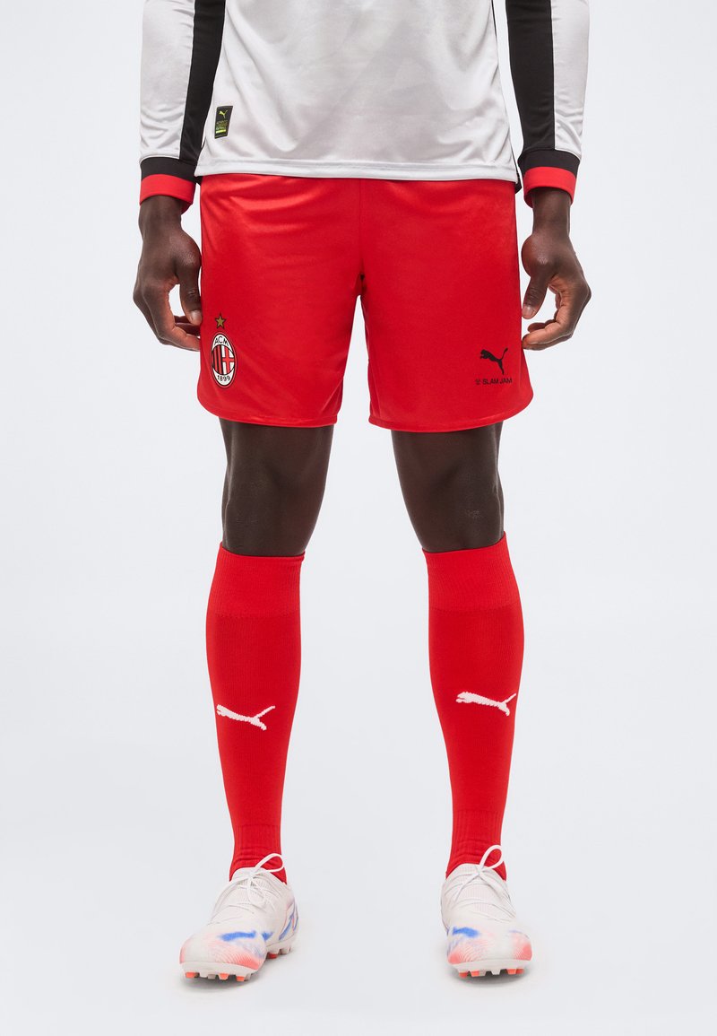 Άτομο που φορά κόκκινα ποδοσφαιρικά σορτς AC Milan, κόκκινες κάλτσες Puma, λευκά παπούτσια με μπλε και πορτοκαλί λεπτομέρειες και λευκό μακρυμάνικο μπλουζάκι με μαύρες ρίγες.