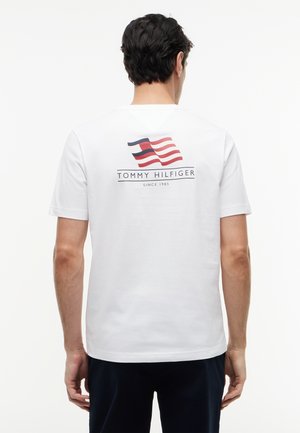 Mand iført hvid Tommy Hilfiger t-shirt med rødt, hvidt og blåt flaglogo og teksten "Tommy Hilfiger Since 1985" på bagsiden.