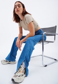 Top beige ajustado, jeans claros de campana con aberturas laterales y zapatillas gris y blancas. El modelo se sienta en una silla de cromo frente a un fondo blanco.