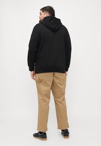 Zwarte hoodie met lange mouwen, gecombineerd met beige broek. De outfit heeft een relaxte pasvorm en zichtbare zakken op de broek.