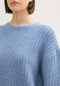 Hellblau gestrickter Pullover mit gerippter Textur, rundem Ausschnitt und überschnittenen Schultern, der ein dezentes Schimmern im gesamten Material aufweist.