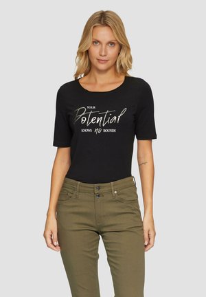 Schwarzes T-Shirt mit kurzen Ärmeln und dem Spruch "DEIN POTENZIAL KENNT KEINE GRENZEN" in metallicfarbener Schrift. Kombiniert mit olivgrünen Hosen.