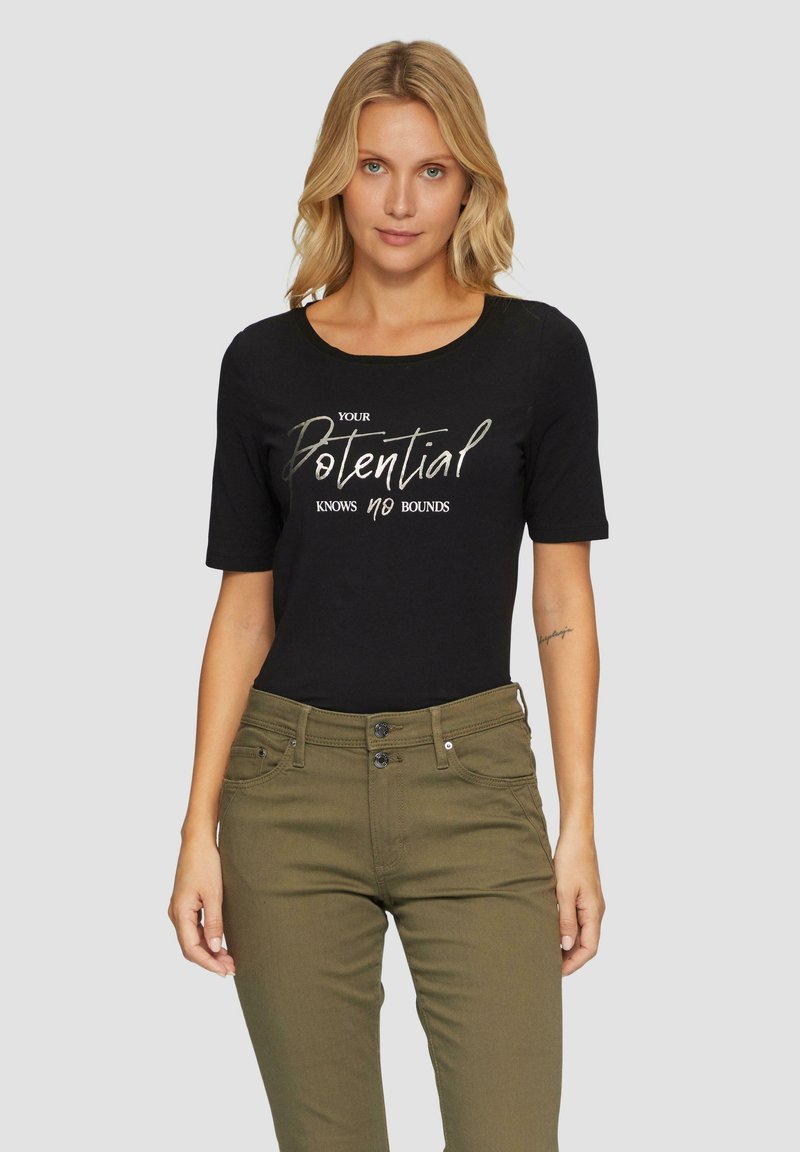 Zwart t-shirt met korte mouwen met de tekst "JE POTENTIE KENT GEEN BEOEFENING" in metallic letters. Genoemd met olijfgroene broek.