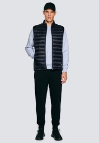 Jonge man met zwarte pet, zwart doorgestikt vest over een lichtgrijze trui, zwarte joggingsbroek en zwarte sneakers, staand met één hand in de broekzak.