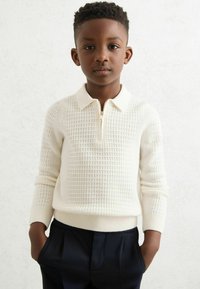 Biely zipsveter s textúrovaným štvorcovým vzorom, s golierom a rebrovanými manžetami, v kombinácii s tmavomodrými nohavicami.