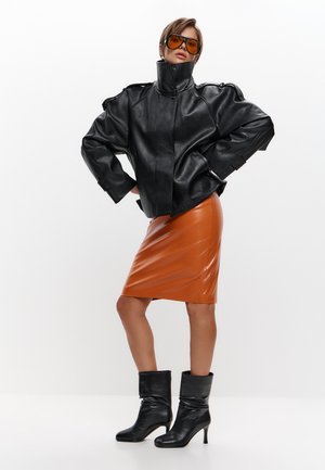 Frau trägt übergroße schwarze Lederjacke, orangefarbenen Lederrock, schwarze Stiefel mit Absatz und bernsteinfarbene Brille, posiert mit Händen in die Hüften gestemmt.