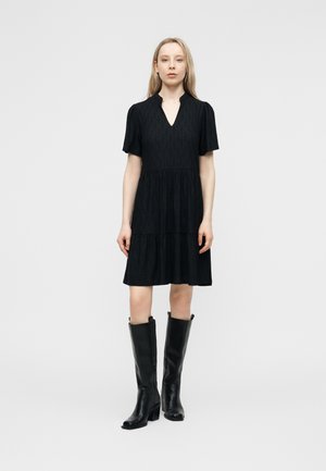 Kobieta ubrana w czarną sukienkę o linii A z krótkim rękawem i subtelnym wzorem, w zestawieniu z czarnymi skórzanymi kozakami za kolano, stojąca na białym tle.