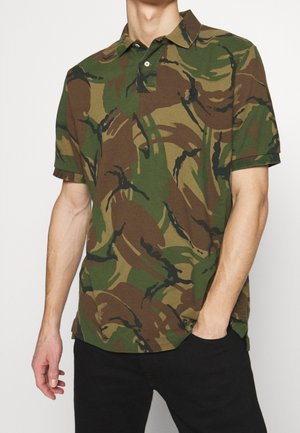 Man die een groen-bruine camouflageprint korte mouwen polo draagt met zwarte broek, één hand in de zak, tegen een effen achtergrond.