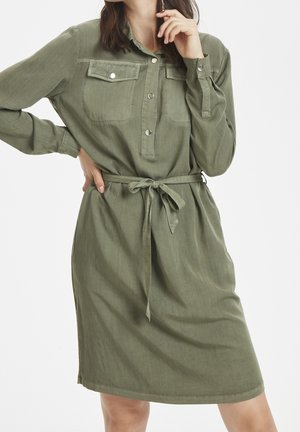 Femme portant une robe-chemise ceinturée de couleur vert olive à manches longues, avec des poches avant et des boutons-pression, posant avec une main sur la hanche et l'autre près du visage.