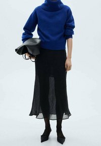 Kobieta w niebieskim swetrze z golfem, przezroczystej czarnej spódnicy midi, czarnych rajstopach i czarnych szpilkach, trzymająca czarną skórzaną torbę.