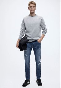 Šedá mikina, modré ošoupané džíny a černé tenisky. Model drží černou tašku, která představuje neformální styl a uvolněný střih.