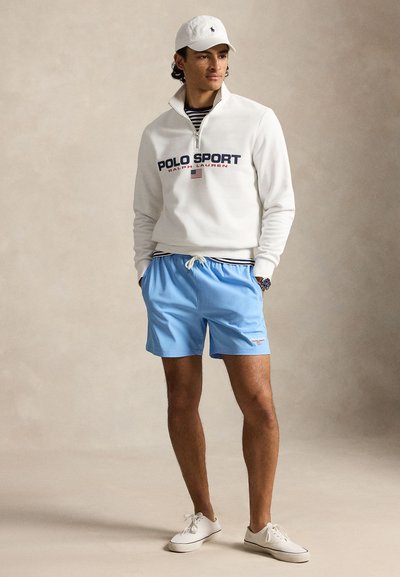 Mees, kes kannab valget Polo Sporti neljandiku tõmbega dressipluusi, helesiniseid nööriga lühikesi pükse, valget nokamütsi ja valgeid tenniseid, seisab käed taskutes.