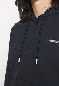 Sudadera con cremallera en azul marino, hecha de una mezcla de algodón. Cuenta con capucha ajustable, cremallera frontal y un logo blanco de Calvin Klein en el pecho.