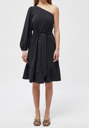 Femme portant une robe noire asymétrique à une épaule avec une manche longue bouffante sur le bras gauche, ceinture à la taille, ourlet à volants et talons ouverts couleur olive.