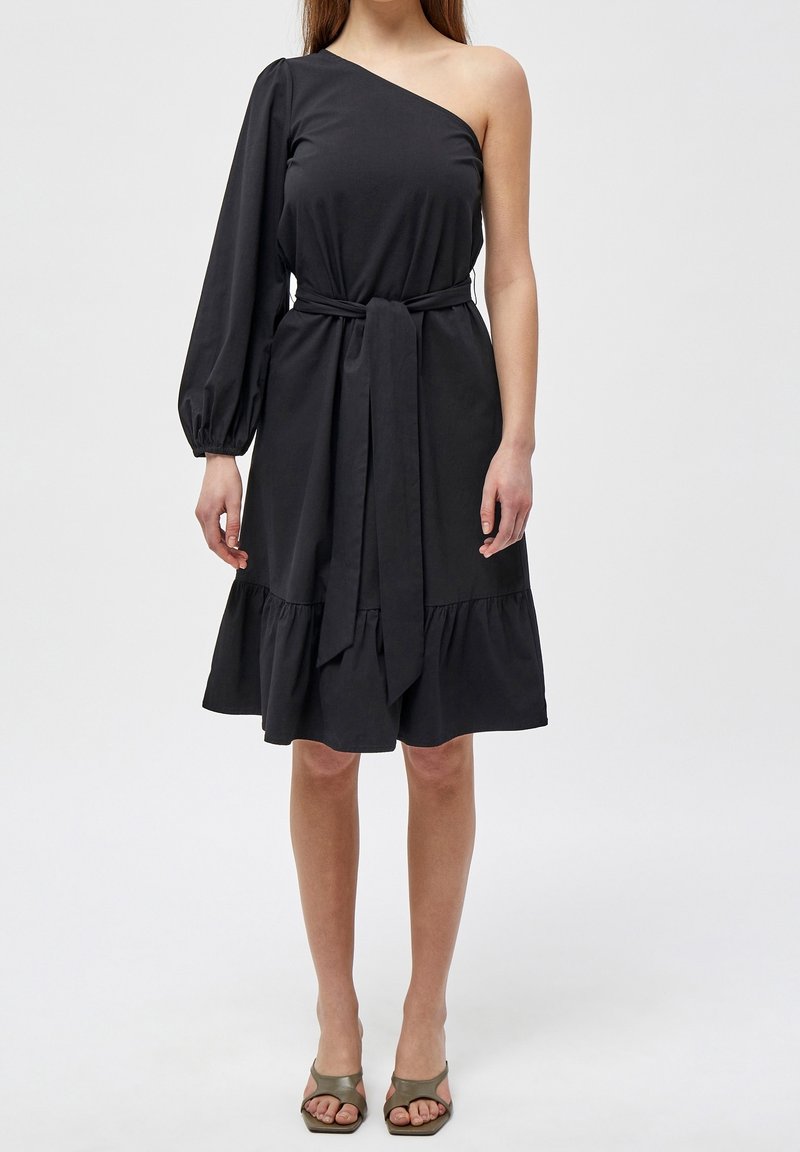 Femme portant une robe noire asymétrique à une épaule avec une manche longue bouffante sur le bras gauche, ceinture à la taille, ourlet à volants et talons ouverts couleur olive.
