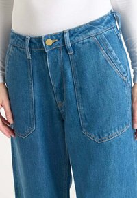 Pantalones de mezclilla azul claro con corte holgado, que cuentan con bolsillos frontales, un cierre con botón dorado y costuras de contraste.