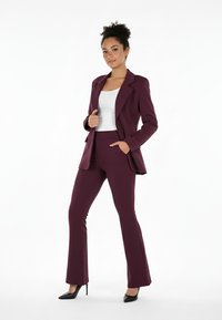 Bordowy garnitur z dopasowaną marynarką na dwa guziki i rozszerzanymi spodniami, zestawiony z białą bluzką i czarnymi szpilkami. Gładka faktura tkaniny.