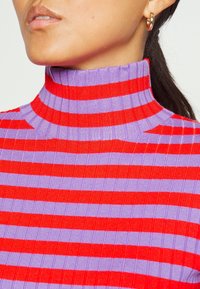 Nära bild av en person som bär en ribbad polotröja med växelvis röda och lila horisontella ränder samt en gul hoop-örhänge.