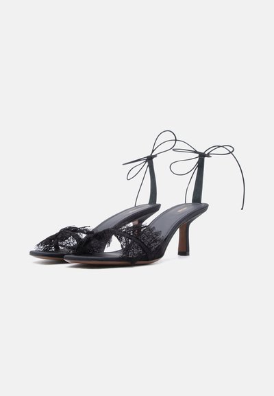 Sandales à talons hauts noires avec détail en dentelle sur la bride avant et fines brides à la cheville nouées en nœuds sur fond blanc.