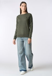 Maglione verde oliva a maniche lunghe con scollo rotondo e logo ricamato. Abbinato a jeans a gamba larga blu chiaro e sneakers bianche.