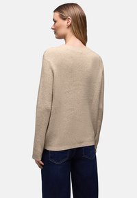Femme de profil portant un pull en maille beige texturé et un jean bleu foncé sur un fond clair uni.