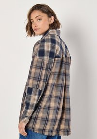 Plaid shirt in tan and navy, featuring a relaxed fit, collar, and frayed hem. Made of soft fabric with a textured plaid pattern.