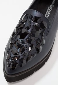 Černá kožená slip-on obuv s texturovaným povrchem zdobeným velkými černými kameny, zužující špičkou a texturovanou černou podrážkou.