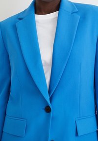 Blazer bleu vif en tissu texturé, avec un col cranté, un bouton noir unique et deux poches latérales. T-shirt blanc en dessous.