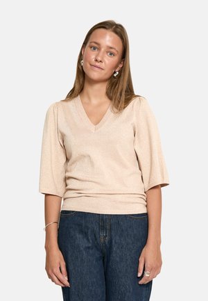 Jauna moteris su tiesiais šviesiai rudos spalvos plaukais, dėvinti smėlio spalvos V formos iškirptės megztinį su alkūnių ilgio rankovėmis ir tamsiai mėlynas džinsines kelnes.