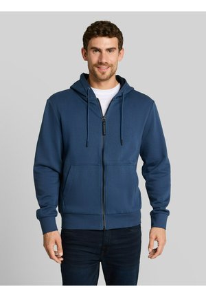 Uomo che indossa una felpa con cappuccio blu navy con cerniera sopra una camicia bianca, in piedi davanti a uno sfondo grigio chiaro uniforme, sorridente con le mani rilassate.