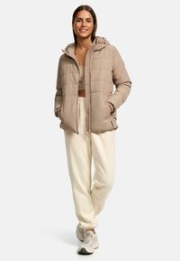 Beige gewatteerde jas met capuchon, voorzien van een gequilt ontwerp, voorkantzakken en rits sluiting, gecombineerd met crèmekleurige joggingbroek en sneakers.