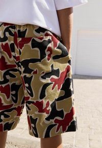 Camo shorts in kaki, rood, zwart en crème met een zak op de achterkant. De stof is katoen met een gladde textuur en een casual pasvorm.