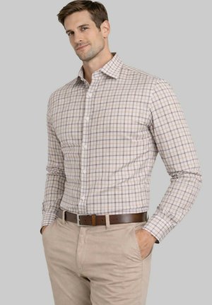 Homme portant une chemise à manches longues beige et marron à carreaux, rentrée dans un pantalon beige clair avec une ceinture marron, les mains dans les poches.