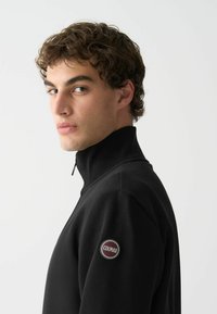 Fekete, félig cipzáros pulóver, puha anyagból készült. Magas gallérral rendelkezik, és egy kerek Colmar logó folt található a bal válltömjén.