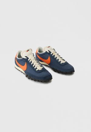 Par navyblå og orange Nike løbesko med hvide snørebånd og teksturerede sorte såler på en ensfarvet lys baggrund.